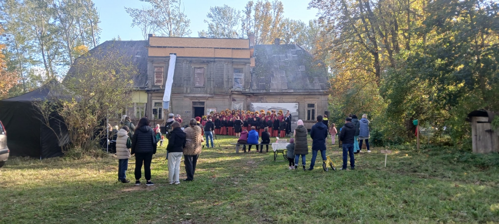 Attēlā redzama Ālaves kungu māja no attāluma, pie mājas notiek pasākums un priekšplānā redzamas skatītāju muguras.