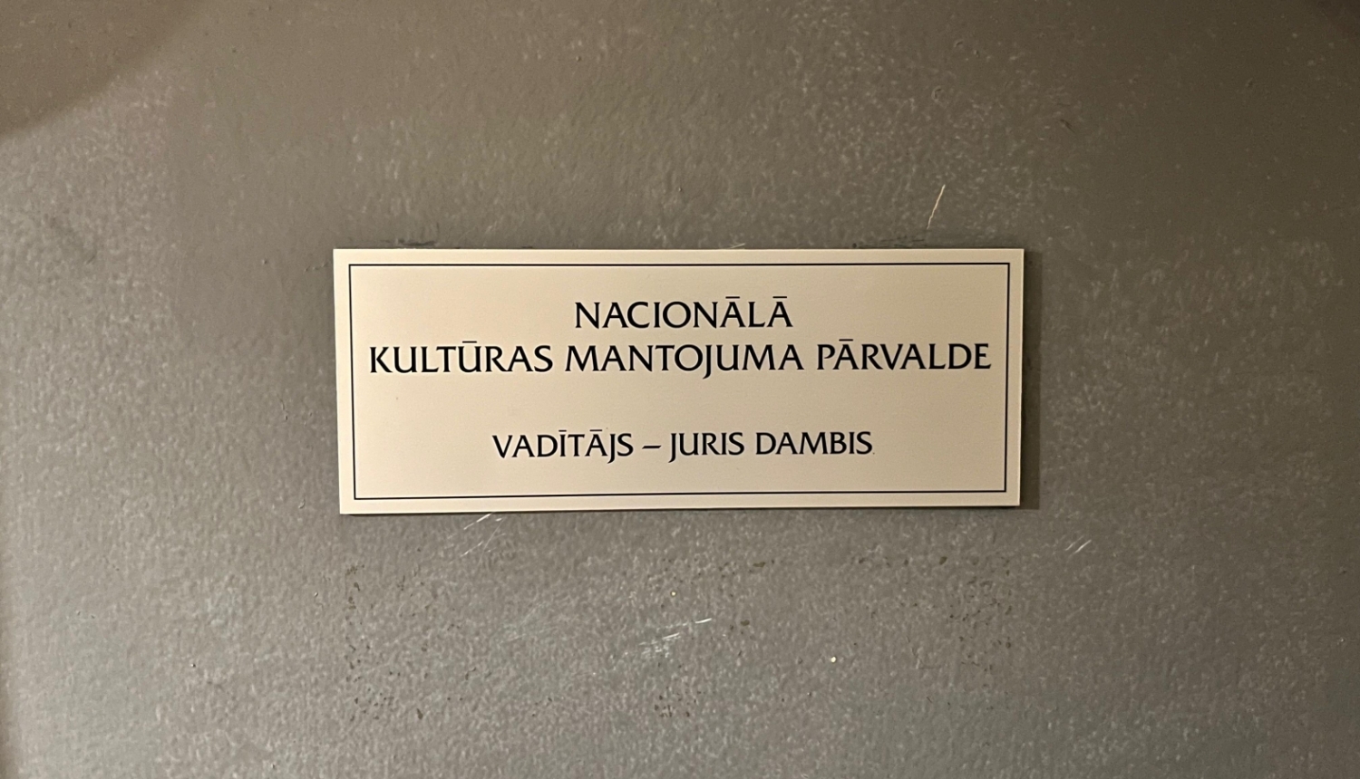 Plāksnīte pie Nacionālās kultūras mantojuma pārvaldes vadītāja durvīm, kas saka "Nacionālā kultūras mantojuma pārvalde"; vadītājs - Juris Dambis. Krāsotās koka durvis ir pelēcīgas, plāksne četrkantīga, neliela, metāla ar melniem burtiem.